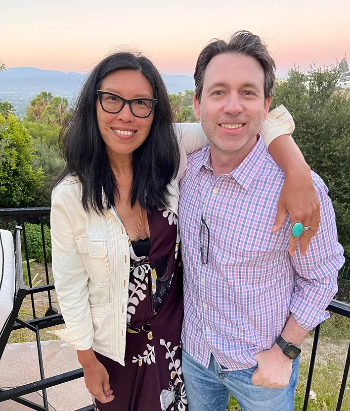 Smiling woman with glasses and man posing outdoors on a patio with greenery and mountains in background.